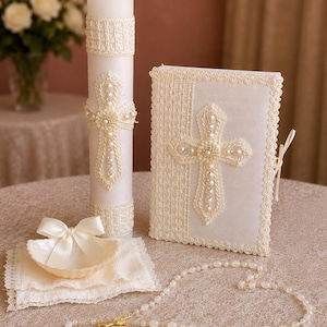 May include: A white ceremonial set featuring a tall pillar candle, a rectangular book, a small shell-shaped dish with a satin bow, and a beaded rosary. The candle and book are decorated with cream-colored lace trim, braided rope, and faux pearls.