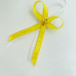 May include: A yellow ribbon with gold glitter trim tied into a bow with a gold cross charm. The ribbon has the text "Kuku Freeman 05-10-16 + 07-08-23" printed on it.