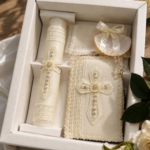 May include: A white gift box containing a cream-colored candle wrapped in lace and pearls, a matching lace-covered book with a pearl cross embellishment, a small white shell with a satin ribbon bow, and a delicate pearl rosary with a gold cross.