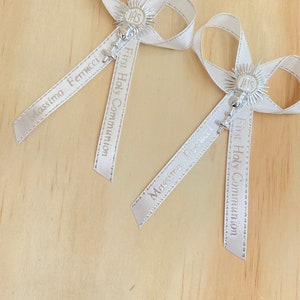 May include: Two white satin ribbon bows with silver charms and the text "First Holy Communion" and names "Massimo Fenucci" and "Mirco Brunelli".