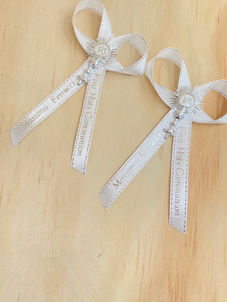 May include: Two white satin ribbon bows with silver charms and the text "First Holy Communion" and names "Massimo Fenucci" and "Mirco Brunelli".