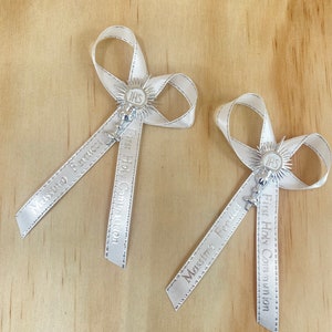 May include: Two white satin bows with silver lettering and a silver charm. The bows are tied with a silver ribbon and have the text "First Holy Communion" and "Massimo Ferrucci" on them.
