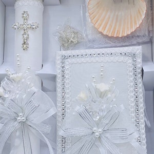 May include: A white baptism set with a candle, a shell, and a small white book with a silver bow. The candle has a silver cross with rhinestones and a white bow with pearls.