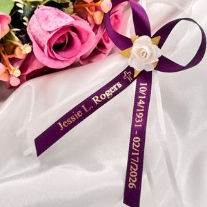 May include: A purple ribbon with gold text reading "Jessie L. Rogers" and dates. A white flower and cross accent the ribbon. Pink roses and white fabric are in the background.