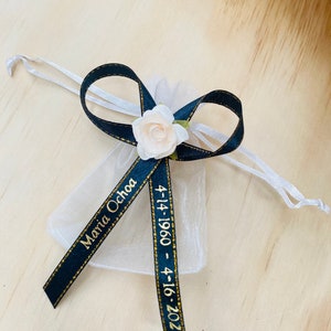 May include: A white organza bag with a black ribbon tied in a bow. The ribbon has gold lettering that reads "Maria Ochoa 4-14-1960 - 4-16-2023".