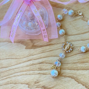 K&ouml;nnte beinhalten: Eine rosa Organzabtasche mit einem goldenen Band, auf dem "Mi Bautizo 8-7-21" und "Andrea's First Holy Communion" steht. Neben der Tasche liegt ein wei&szlig;es Perlenarmband mit einem goldenen Anh&auml;nger der Jungfrau Maria und einem goldenen Engelsanh&auml;nger.