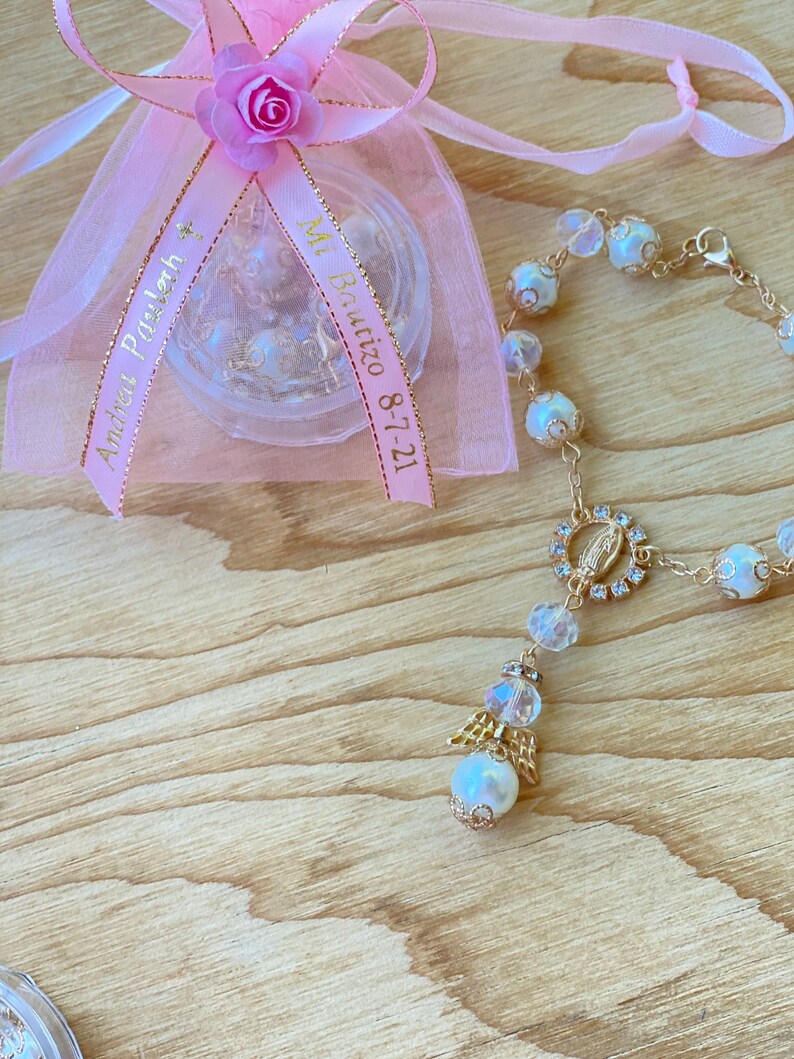 K&ouml;nnte beinhalten: Eine rosa Organzabtasche mit einem goldenen Band, auf dem "Mi Bautizo 8-7-21" und "Andrea's First Holy Communion" steht. Neben der Tasche liegt ein wei&szlig;es Perlenarmband mit einem goldenen Anh&auml;nger der Jungfrau Maria und einem goldenen Engelsanh&auml;nger.