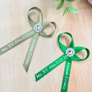 May include: Two green and white satin ribbon favors with a rhinestone embellishment. The ribbons are tied in a bow and have the text "Mis XV Años 11-23-24 Amayah Estrada" printed on them.