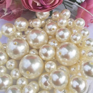 May include: A collection of ivory-colored, round beads of varying sizes, with a glossy finish. The beads are arranged in a clear glass bowl, with pink fabric flowers in the background. The beads have small holes.