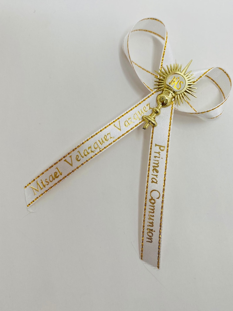 May include: White satin ribbon with gold trim and text that reads "Primera Comuni&oacute;n" and "Misael Vel&aacute;zquez V&aacute;zquez". A gold chalice with a sunburst design and the letters "IHS" is attached to the ribbon.