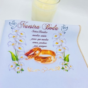 May include: White fabric with gold trim and a wedding theme. The fabric features a floral design with white calla lilies, two gold wedding bands, and two white doves. The text reads "Nuestra Boda" and "Señor Bendice nuestra unión para que nuestro amor perdure por siempre."