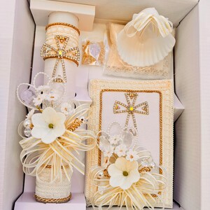 May include: A white baptism set with a candle, a shell, a prayer book, and a small white flower arrangement. The candle is decorated with rhinestones and a gold cross. The prayer book is also decorated with a gold cross and rhinestones.