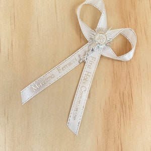 May include: White satin ribbon with the words "First Holy Communion" and a name printed on it. The ribbon is tied into a bow with a silver charm in the centre. The charm features a chalice with a host and the letters "IHS" above it.