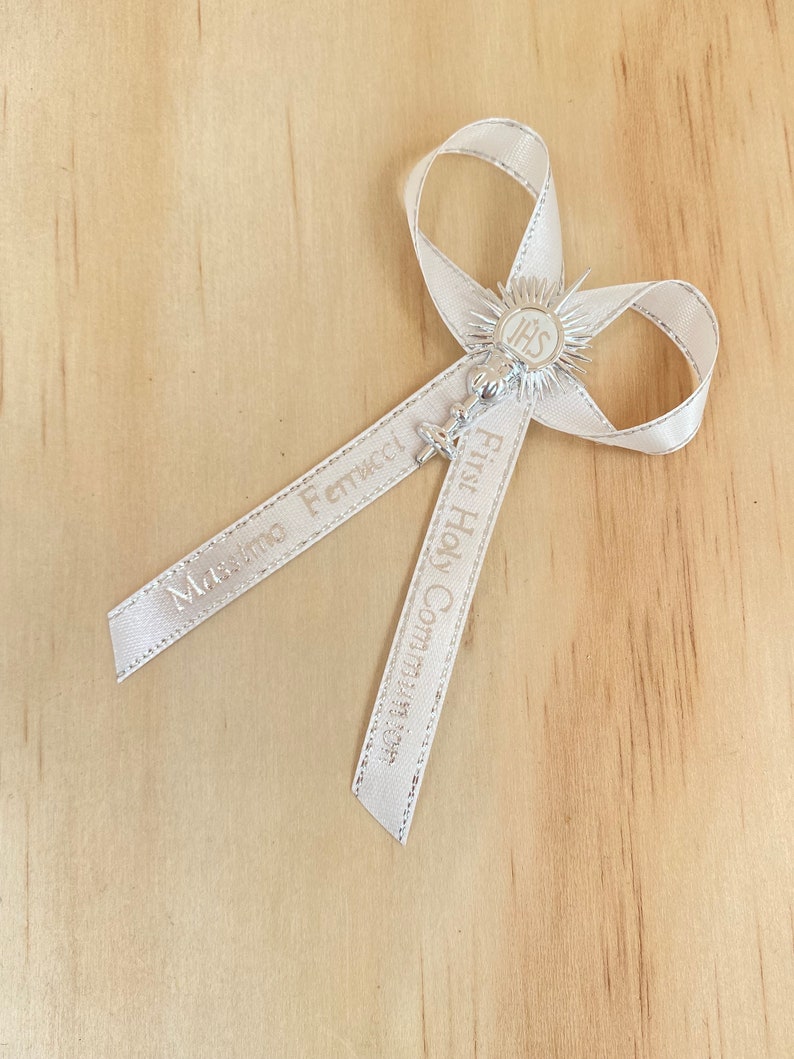 May include: White satin ribbon with the words "First Holy Communion" and a name printed on it. The ribbon is tied into a bow with a silver charm in the centre. The charm features a chalice with a host and the letters "IHS" above it.