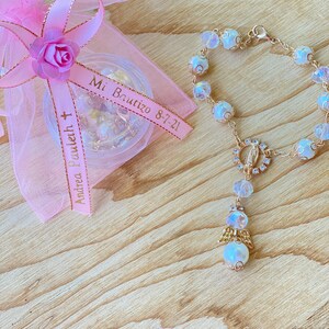 K&ouml;nnte beinhalten: Ein wei&szlig;es und goldenes Perlenarmband mit einem kleinen Engelsanh&auml;nger und einem gr&ouml;&szlig;eren Anh&auml;nger mit einer Darstellung der Jungfrau Maria. Das Armband liegt auf einer Holzoberfl&auml;che mit einer rosa Organzas&auml;ckchen mit einem Band, auf dem "Mi Bautizo 8-7-21" und "Andrea Bouquet" steht.