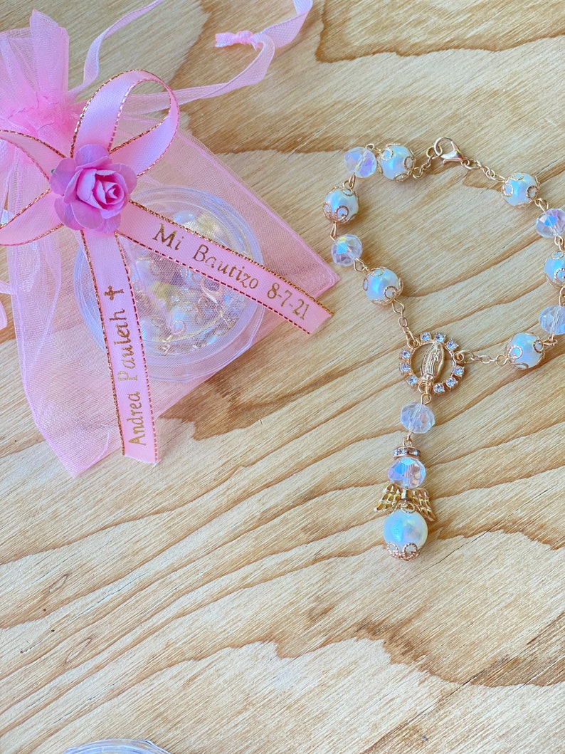K&ouml;nnte beinhalten: Ein wei&szlig;es und goldenes Perlenarmband mit einem kleinen Engelsanh&auml;nger und einem gr&ouml;&szlig;eren Anh&auml;nger mit einer Darstellung der Jungfrau Maria. Das Armband liegt auf einer Holzoberfl&auml;che mit einer rosa Organzas&auml;ckchen mit einem Band, auf dem "Mi Bautizo 8-7-21" und "Andrea Bouquet" steht.