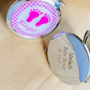 May include: Two silver compact mirrors. One mirror has a pink and white gingham pattern with pink footprints and the text "It's a girl!" The other mirror has the text "Maribel's Baby Shower 6-15-22".