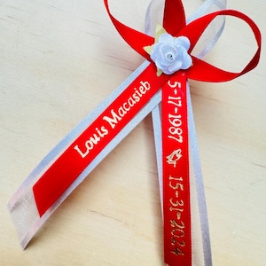 May include: Red and white ribbon with a white flower and gold lettering. The ribbon says "Louis Macasieb 5-17-1987  15-31-2024".