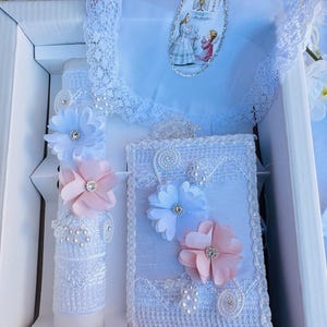 May include: A white box containing a set of baptism or communion items. The set includes a white candle with a lace cover and pink and blue flowers, a white pillow with lace trim and pink and blue flowers, and a blue handkerchief with lace trim and a religious image.