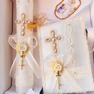 May include: A white religious set including a decorated candle, a book, and a prayer card. The candle and book feature jeweled crosses and gold-colored chalice emblems. The prayer card has a gold border and an illustration of two figures.