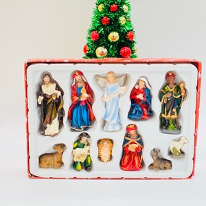 May include: A Christmas nativity set with figurines of biblical figures, including Mary, Joseph, and the Three Wise Men, arranged in a red box. A small Christmas tree with a red star on top is behind the box.