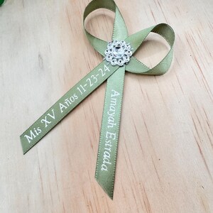 May include: A green satin ribbon with a silver rhinestone embellishment. The ribbon is tied in a bow and has the text "Mis XV Años 11-23-24" and "Amayah Estrada" printed on it.