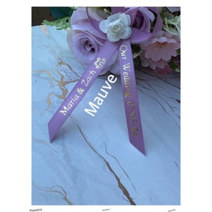 May include: Purple satin ribbon with the text "Our Wedding 07-05-24" and "Maria & Zach" in gold lettering. The ribbon is tied around a bouquet of purple and pink flowers.