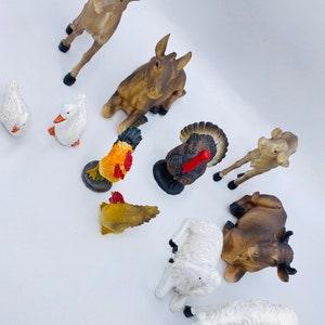 May include: A collection of farm animal figurines, including a brown cow, a white sheep, a rooster, a turkey, and a brown and white goat. The figurines are made of painted plastic and are about 3 inches tall.