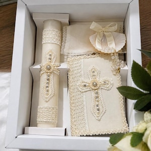 May include: A boxed set of religious items, including a candle, book, and shell-shaped accessory. The items are cream-colored with lace and pearl embellishments. The candle and book feature cross designs. The set is presented in a white box.