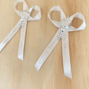 May include: Two white satin ribbon bows with silver charms and the text "First Holy Communion" and "Massimo Bertucci" printed on the ribbons.