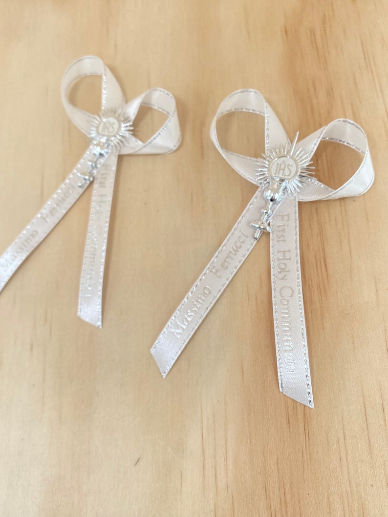 May include: Two white satin ribbon bows with silver charms and the text "First Holy Communion" and "Massimo Bertucci" printed on the ribbons.