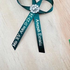 May include: A dark green ribbon bow with a sparkling rhinestone centerpiece. The ribbon tails are inscribed with the text "Mis XV Años 11-23-24" and "Amayah Estrada" in white lettering. The bow is displayed on a light wood surface.