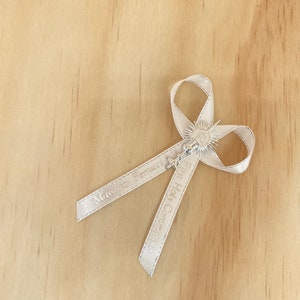 May include: White satin ribbon with a silver charm featuring a chalice and host. The ribbon is tied in a bow and has the text "Massimo Ferraro" and "First Holy Communion" printed on it.