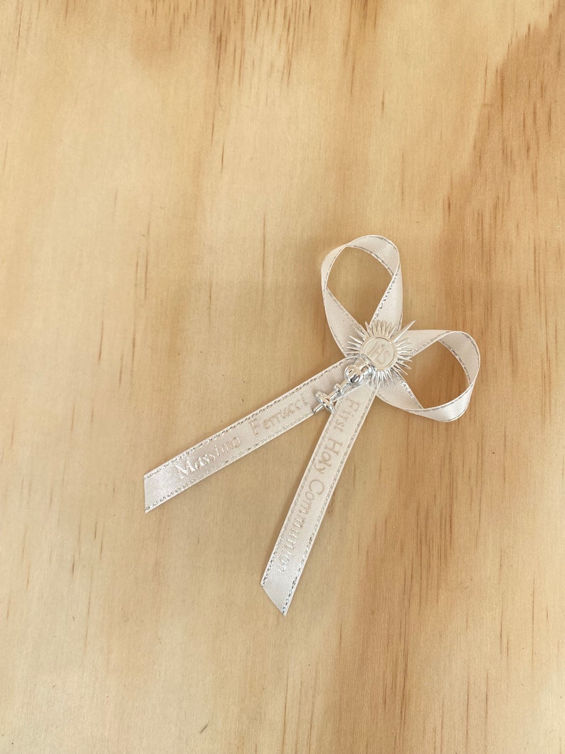 May include: White satin ribbon with a silver charm featuring a chalice and host. The ribbon is tied in a bow and has the text "Massimo Ferraro" and "First Holy Communion" printed on it.