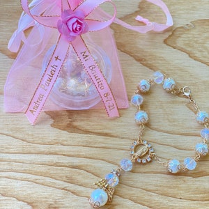 K&ouml;nnte beinhalten: Eine rosa Organzabtasche mit einem goldenen Band, auf dem "Mi Bautizo 8-7-21" und "Andrea Pauleth" steht, ist darum gebunden. Ein wei&szlig;es Perlenarmband mit einer goldenen Kette und einem Anh&auml;nger der Jungfrau Maria liegt neben der Tasche.