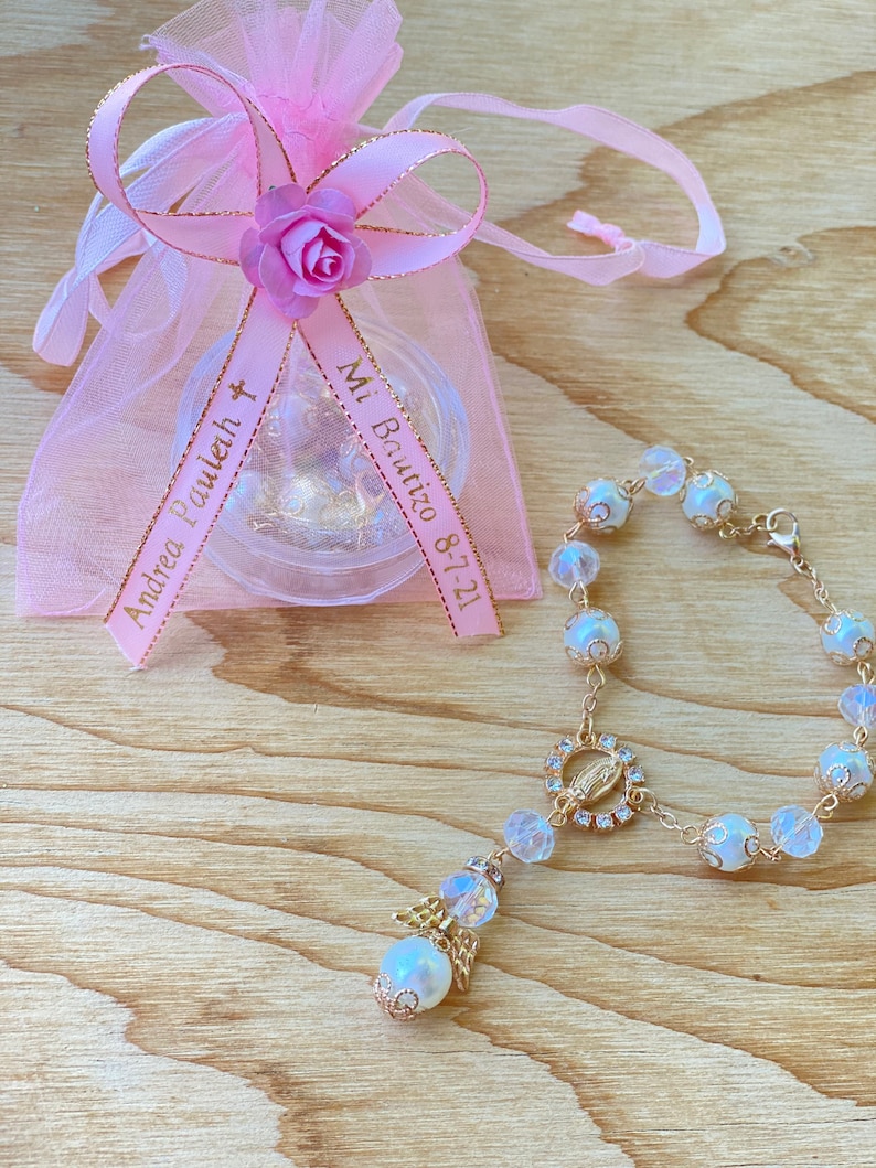 K&ouml;nnte beinhalten: Eine rosa Organzabtasche mit einem goldenen Band, auf dem "Mi Bautizo 8-7-21" und "Andrea Pauleth" steht, ist darum gebunden. Ein wei&szlig;es Perlenarmband mit einer goldenen Kette und einem Anh&auml;nger der Jungfrau Maria liegt neben der Tasche.