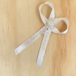 May include: A white satin ribbon bow with a silver charm featuring a chalice and the inscription "IHS". The ribbon is printed with the text "Massimo Ferrucci" and "Prima Comunione".