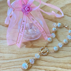 K&ouml;nnte beinhalten: Eine wei&szlig;e und goldene Rosenkranz mit einem klaren Kristall-Anh&auml;nger und einem kleinen Engels-Anh&auml;nger. Der Rosenkranz befindet sich in einem rosa Organza-Beutel mit einem goldenen Band, auf dem "Andrea Pauleh" und "Mi Bautizo 8-7-21" steht.