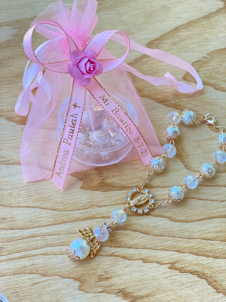 K&ouml;nnte beinhalten: Eine wei&szlig;e und goldene Rosenkranz mit einem klaren Kristall-Anh&auml;nger und einem kleinen Engels-Anh&auml;nger. Der Rosenkranz befindet sich in einem rosa Organza-Beutel mit einem goldenen Band, auf dem "Andrea Pauleh" und "Mi Bautizo 8-7-21" steht.