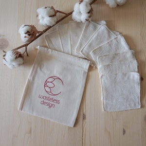 May include: A set of white reusable facial rounds and a matching drawstring bag with the text "wasteless design" in a burgundy color. The rounds are arranged on a light wooden surface, with cotton branch accents.