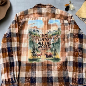 Könnte beinhalten: Ein braunes und weißes kariertes Flanellhemd mit einem gebleichten Effekt. Die Rückseite des Hemdes zeigt ein Aquarellbild einer Waldszene mit einem Reh und einem Bären. Der Text "GO INTO ALL THE WORLD. - MARK 16:15" ist auf dem Hemd aufgedruckt.