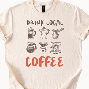 Peut inclure: Un t-shirt beige avec le texte "DRINK LOCAL COFFEE" en noir et rouge. Le t-shirt présente des illustrations de différentes méthodes de préparation du café, notamment une presse française, une cafetière, un filtre à café et une machine à espresso.