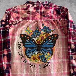 Könnte beinhalten: Ein rosafarbenes und schwarzes kariertes Flanellhemd mit einem gebleichten Effekt. Das Hemd zeigt ein Motiv eines blauen Schmetterlings mit dem Text "Live Life in Full Bloom".