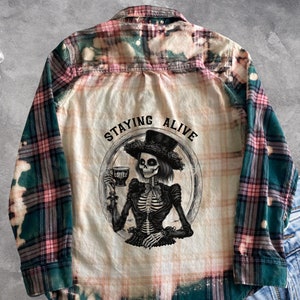 Puede incluir: Una camisa de franela a cuadros verde y rosa con efecto blanqueado. La camisa presenta un gráfico de un esqueleto sosteniendo una copa de vino con el texto "Staying Alive".