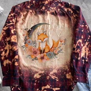 Könnte beinhalten: Ein weinrotes und cremefarbenes Button-Down-Shirt mit gebleichtem Design. Auf der Rückseite des Hemdes befindet sich eine Aquarellillustration eines Fuchses, der auf einem Mond sitzt und von Blumen umgeben ist. Das Hemd wird mit weißem und blauem Denim präsentiert.