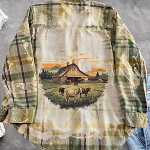 May include: A green and yellow plaid flannel shirt with a bleached design of a farm scene with a barn, cows, and mountains. The shirt has a faded look and is perfect for a casual, rustic style.