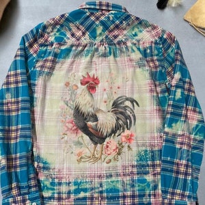 Puede incluir: Una camisa de franela a cuadros azules y blancos con un diseño blanqueado que presenta un gallo y flores en la espalda.