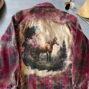 May include: A red and black checked shirt with a distressed finish. The back of the shirt displays a circular image of a brown horse in a forest. The shirt has long sleeves and a standard collar.