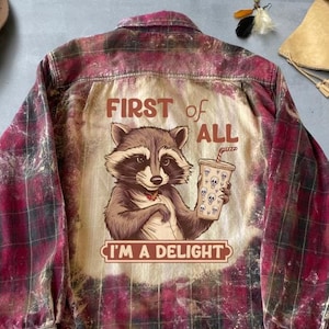 Może przedstawiać: Czerwono-czarna koszula w kratę z flaneli z efektem postarzenia. Z tyłu znajduje się grafika szopa trzymającego kubek ozdobiony czaszkami, z napisem "FIRST of ALL I'M A DELIGHT".