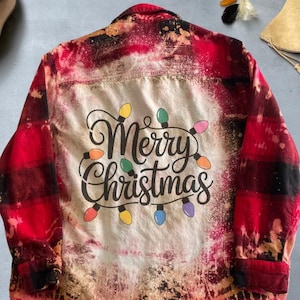 May include: Red and black plaid button-down shirt with a bleached design. The back of the shirt features the words "Merry Christmas" in a script font, surrounded by colorful Christmas lights. The shirt has long sleeves.